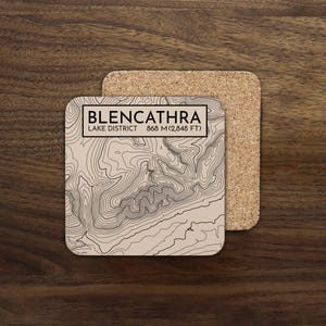 May include: Square coaster featuring a topographic map of Blencathra in the Lake District. The beige coaster has black contour lines and text: "BLENCATHRA LAKE DISTRICT 868 M (2,848 FT)". A second cork coaster is visible behind it.