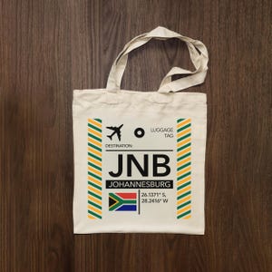 Peut inclure: Un sac fourre-tout en toile couleur crème arborant un motif graphique rappelant une étiquette de bagage pour Johannesburg, en Afrique du Sud. L'impression inclut le code aéroport JNB, le nom de la ville, le drapeau sud-africain et des coordonnées.