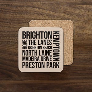 May include: Square coaster featuring a list of Brighton locations in black text, including "BRIGHTON" and "THE LANES". A second cork coaster is behind the first. The coasters are on a wooden surface.