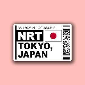 May include: A white sticker with black text and a Japanese flag. The text reads "NRT TOKYO, JAPAN" with coordinates and a barcode. The background is a light pink.