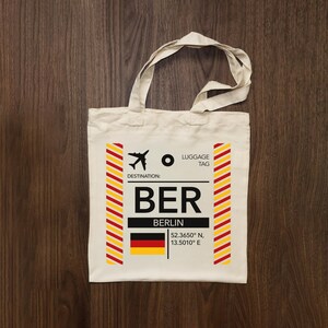 May include: Cream tote bag featuring a Berlin luggage tag design. The design includes the letters "BER", the city name, a plane icon, and the German flag. The bag has two handles and is made of a canvas-like material.