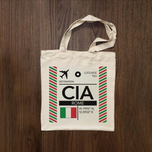 May include: A cream-coloured canvas tote bag featuring a printed graphic resembling an airline luggage tag. The design includes an aeroplane icon, the text LUGGAGE TAG, DESTINATION: CIA, ROME, the Italian flag, and coordinates 41.7992 N, 12.5932 E.