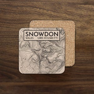 May include: Square coaster with a topographic map design of Snowdon, Wales. The coaster features the text "SNOWDON WALES 1,085 m (3,560 ft)" in a black font. The coaster is tan with black contour lines, and is paired with a cork coaster.