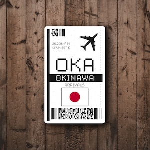 May include: A white airport arrival sign with black text and a red circle on a white background. The sign reads "OKA OKINAWA ARRIVALS" with the coordinates "26.2064° N 127.6465° E" and a small airplane icon.