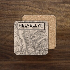 May include: Square coaster with a topographic map of Helvellyn in the Lake District. The coaster has a beige background with black contour lines and the text "HELVELLYN LAKE DISTRICT 950 M (3,116 FT)". A second cork coaster is visible.