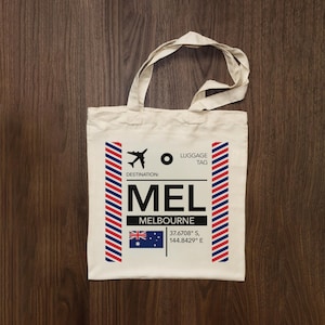 Peut inclure: Un sac fourre-tout en toile couleur crème arborant un motif graphique rappelant une étiquette de bagage d'avion. L'impression comprend une icône d'avion noire, le texte "DESTINATION: MEL MELBOURNE", le drapeau australien et les coordonnées 37.6708° S, 144.8429° E.