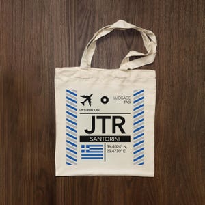 May include: A cream-coloured canvas tote bag featuring a printed luggage tag design. The graphic includes an aeroplane icon, the text LUGGAGE TAG, DESTINATION: JTR, SANTORINI, the Greek flag, and coordinates 36.4024 N, 25.4730 E with blue and grey stripes.
