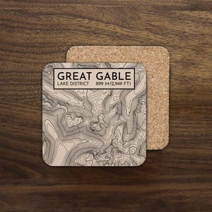May include: Square coaster with a topographic map design. The coaster features the text "GREAT GABLE LAKE DISTRICT 899 M (2,949 FT)" in a rectangular frame. The map is in shades of beige and brown. A second cork coaster is visible.