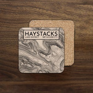 May include: Square coaster featuring a topographic map of Haystacks in the Lake District. The coaster has the text "HAYSTACKS LAKE DISTRICT 597 M (1,959 FT)". A second cork coaster is partially visible.