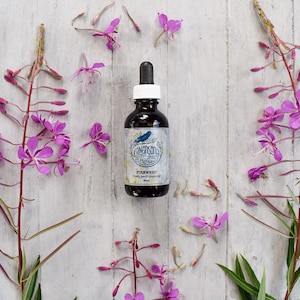 May include: A dark glass bottle with a dropper top and a white label. The label says "RavenSong Herbs" and "Fireweed fresh herb tincture 60ml". The bottle is surrounded by pink flowers on a white wooden background.