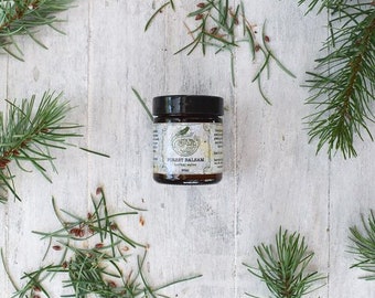 May include: A small glass jar with a black lid and a white label that reads "FOREST BALSAM" with a botanical design. The jar is sitting on a white wooden surface with green pine needles scattered around it.