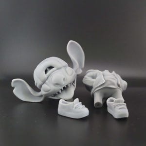 May include: A disassembled gray figurine of a cartoon character, featuring a head with large ears and sharp teeth, a torso, and two small sneakers. The pieces are arranged on a dark gray surface.