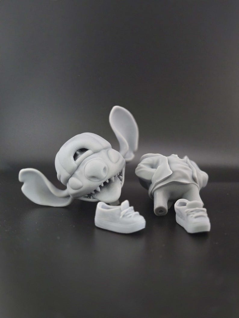 May include: A disassembled gray figurine of a cartoon character, featuring a head with large ears and sharp teeth, a torso, and two small sneakers. The pieces are arranged on a dark gray surface.