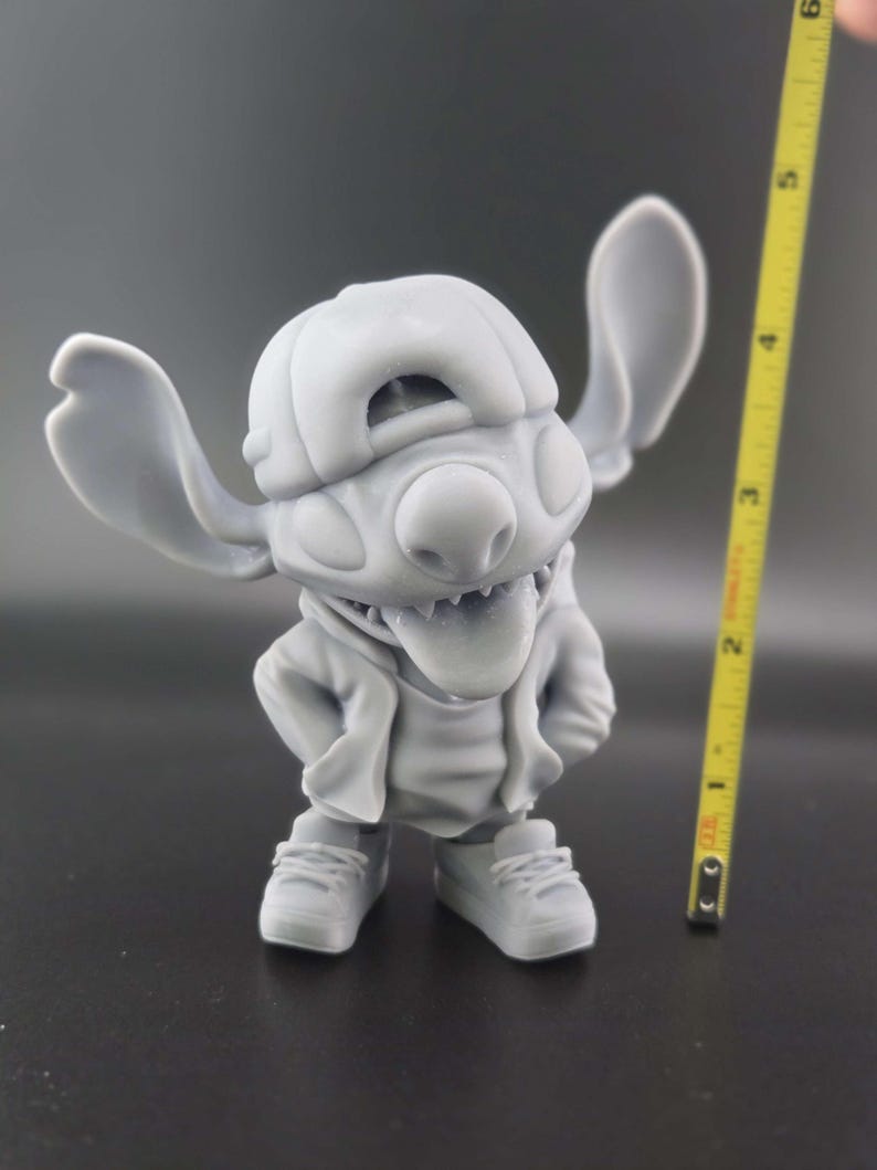 May include: A gray figurine of a cartoon character wearing a backward baseball cap, hoodie, and sneakers. The character has large ears and is sticking out its tongue. A yellow tape measure is next to the figurine, showing a height of approximately 4 inches.