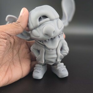 May include: A gray figurine of a cartoon character with large ears, wearing a hat, hoodie, and sneakers. The character has its tongue sticking out and hands in its pockets. The figurine is held by a hand.