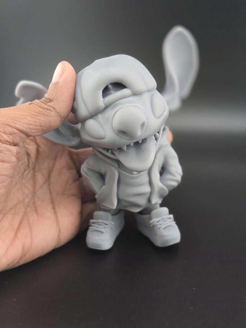 May include: A gray figurine of a cartoon character with large ears, wearing a hat, hoodie, and sneakers. The character has its tongue sticking out and hands in its pockets. The figurine is held by a hand.
