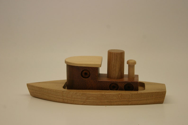 May include: A wooden toy tugboat with a brown hull, a light brown deck, and a light brown smokestack. The tugboat has two round portholes on each side.