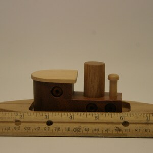 May include: A wooden toy tugboat with a brown hull, a light wood deck, and a single smokestack. The boat is about 6 inches long and is sitting on a wooden ruler.