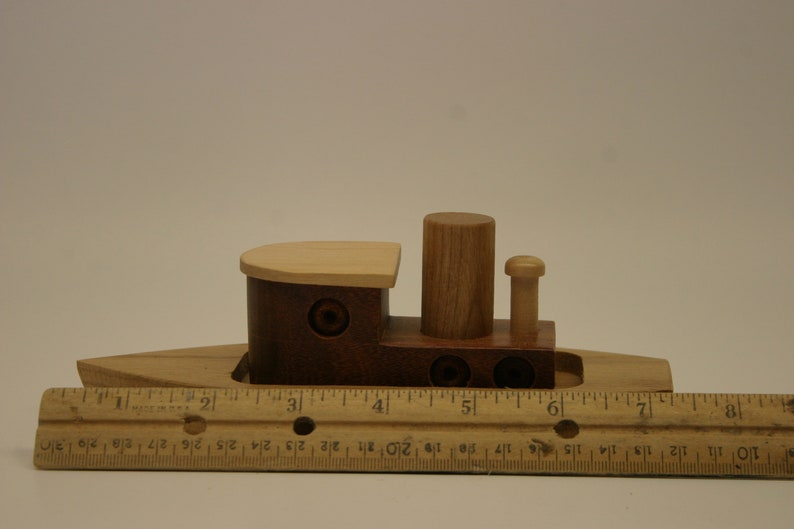 May include: A wooden toy tugboat with a brown hull, a light wood deck, and a single smokestack. The boat is about 6 inches long and is sitting on a wooden ruler.