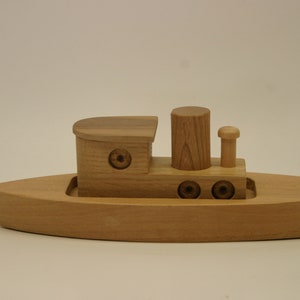 May include: A wooden toy tugboat with a brown stain. The boat has a small cabin, a smokestack, and a small mast with a flag. The boat has three round portholes on each side.