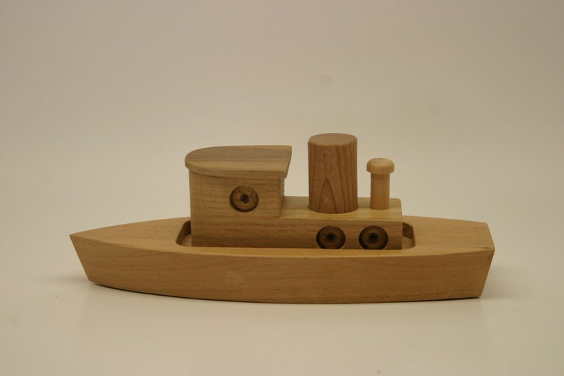 May include: A wooden toy tugboat with a brown stain. The boat has a small cabin, a smokestack, and a small mast with a flag. The boat has three round portholes on each side.