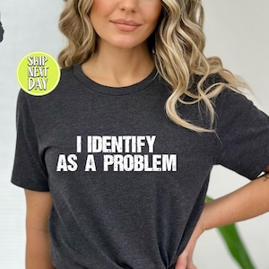 Puede incluir: Camiseta gris oscuro jaspeado con el texto "I Identify As A Problem" en letras blancas. La camiseta es de cuello redondo y la lleva una persona. La imagen también incluye un círculo gris oscuro con las palabras "Dark Grey Heather".