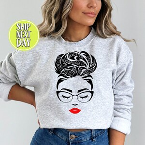 Könnte beinhalten: Grauer Pullover mit einer schwarz-weißen Grafik einer Frau mit einem Dutt. Der Dutt besteht aus Wörtern wie "Queen", "Boss", "Fierce", "Beautiful", "Love", "Magic", "Resilient", "Unapologetic", "Diva" und "Dope".