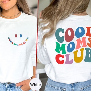 以下が含まれることがあります： レインボーカラーで「COOL MOMS CLUB」と書かれた白い長袖スウェットシャツ。シャツの前側には、同じテキストが小さく書かれたスマイリーフェイスがあります。