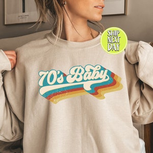 Peut inclure: Un sweat-shirt beige avec un graphisme rétro qui dit "70's Baby" dans des couleurs arc-en-ciel. Le sweat-shirt a une coupe décontractée et un col rond.