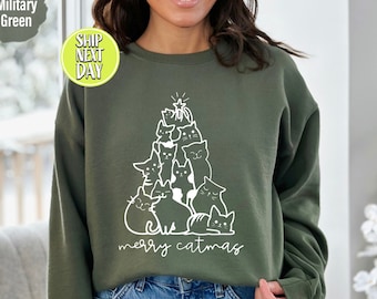 Op de afbeelding: Een groen sweatshirt met een wit ontwerp van een kerstboom gemaakt van katten. De tekst "merry catmas" staat onder de boom.