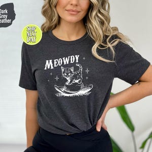 以下が含まれることがあります： 暗いグレーのヘザーTシャツに、カウボーイハットをかぶった猫の白いグラフィックと「MEOWDY」の文字がプリントされています。