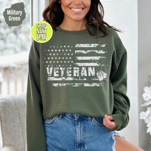 Peut inclure: Un sweat-shirt vert avec un motif de drapeau am&eacute;ricain blanc d&eacute;lav&eacute; et le mot "Veteran" avec des m&eacute;dailles militaires.