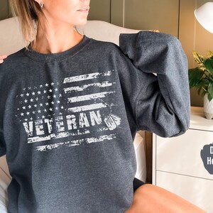 Peut inclure: Un sweat-shirt gris chin&eacute; fonc&eacute; avec un drapeau am&eacute;ricain blanc d&eacute;lav&eacute; et le mot "Veteran" imprim&eacute; dessus.
