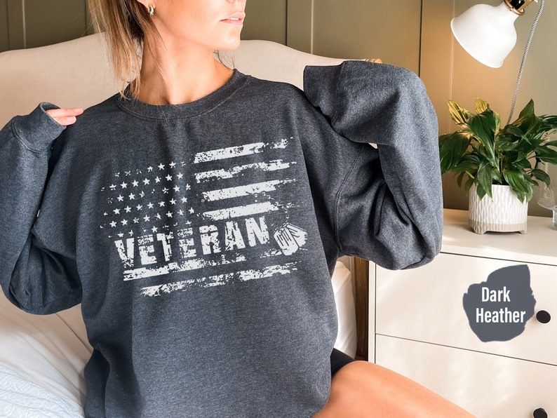 Peut inclure: Un sweat-shirt gris chin&eacute; fonc&eacute; avec un drapeau am&eacute;ricain blanc d&eacute;lav&eacute; et le mot "Veteran" imprim&eacute; dessus.
