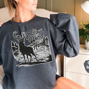 May include: Dark heather gray sweatshirt with a graphic of a deer and the text "Follow the Word Not the Herd" and "Isaiah 8:11". The design is in black and white.