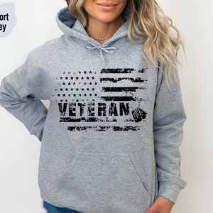 Peut inclure: Un sweat &agrave; capuche gris avec un motif de drapeau am&eacute;ricain us&eacute; et le mot "Veteran" imprim&eacute; en noir. Le drapeau a une silhouette noire d'une plaque d'identit&eacute; militaire.