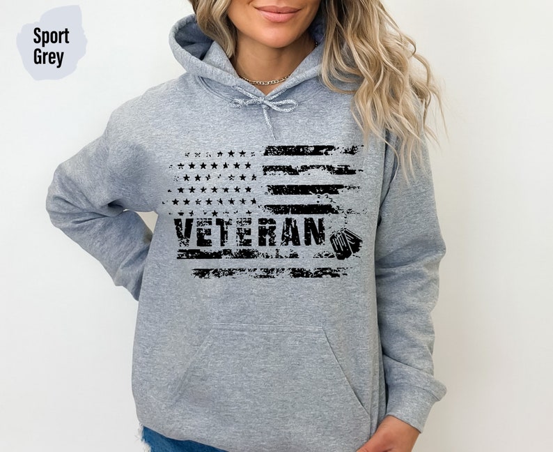 Peut inclure: Un sweat &agrave; capuche gris avec un motif de drapeau am&eacute;ricain us&eacute; et le mot "Veteran" imprim&eacute; en noir. Le drapeau a une silhouette noire d'une plaque d'identit&eacute; militaire.