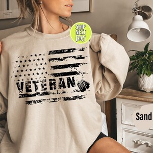 Peut inclure: Un sweat-shirt beige avec un motif de drapeau am&eacute;ricain us&eacute; et le mot "Veteran" en lettres noires.