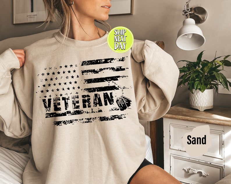 Peut inclure: Un sweat-shirt beige avec un motif de drapeau am&eacute;ricain us&eacute; et le mot "Veteran" en lettres noires.