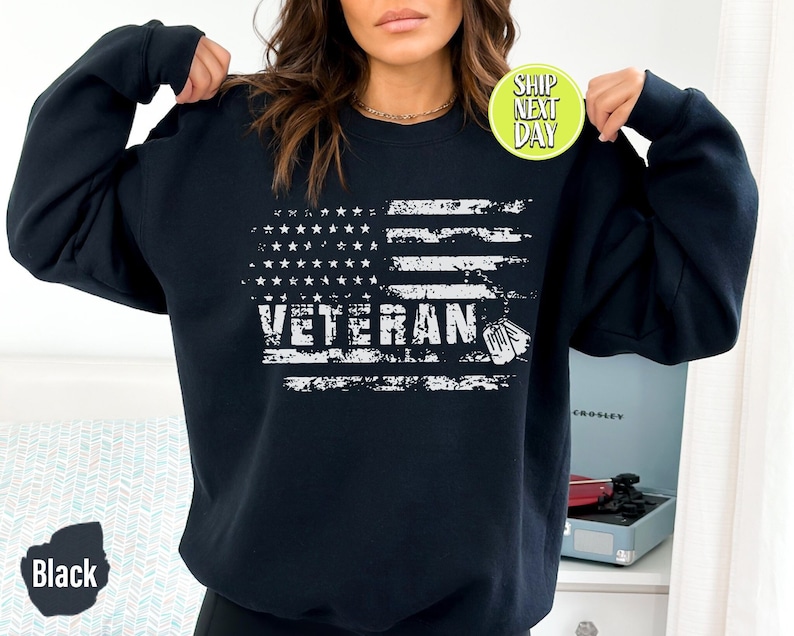Peut inclure: Un sweat-shirt noir avec un drapeau am&eacute;ricain blanc d&eacute;lav&eacute; et le texte "Veteran" avec une petite silhouette blanche d'une plaque d'identit&eacute;.