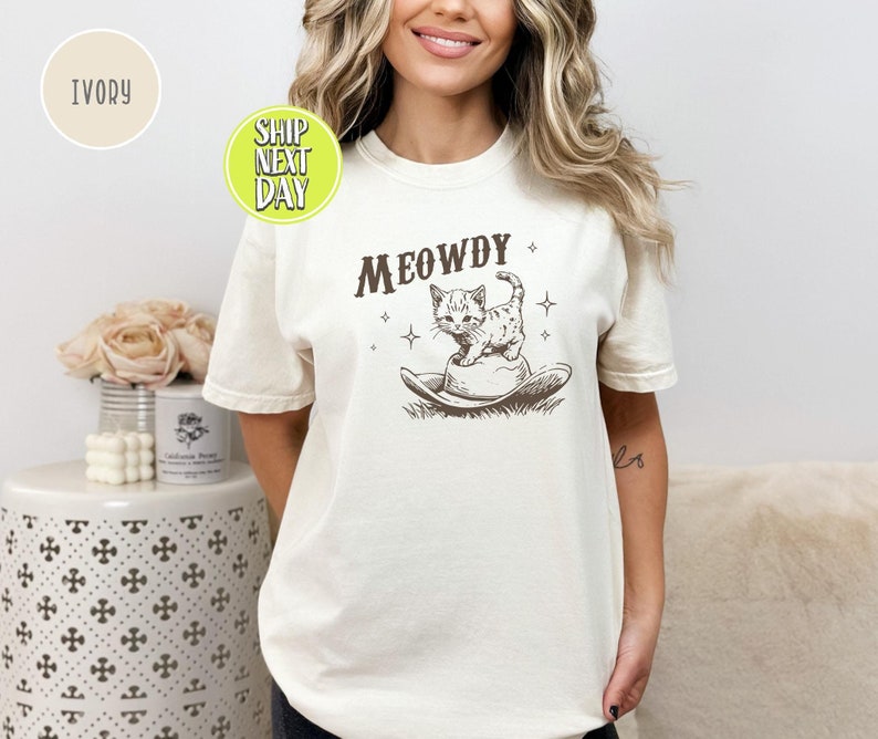 以下が含まれることがあります： アイボリー色のTシャツに、カウボーイハットをかぶった猫のグラフィックと「MEOWDY」の文字がプリントされています。