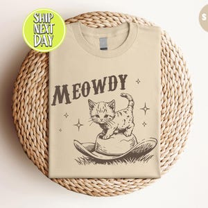 以下が含まれることがあります： ベージュのTシャツに、茶色の「MEOWDY」という文字がプリントされています。カウボーイハットをかぶった漫画の猫が「MEOWDY」の文字の上に座っています。
