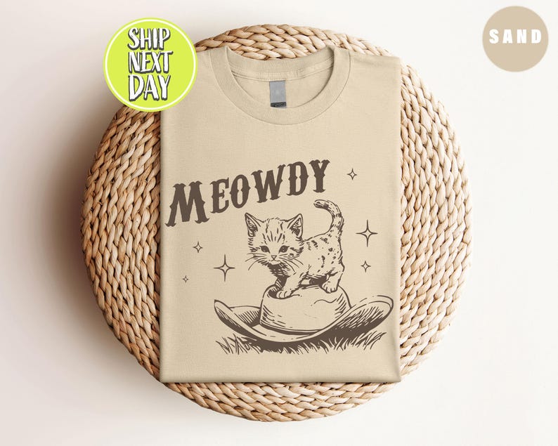 以下が含まれることがあります： ベージュのTシャツに、茶色の「MEOWDY」という文字がプリントされています。カウボーイハットをかぶった漫画の猫が「MEOWDY」の文字の上に座っています。