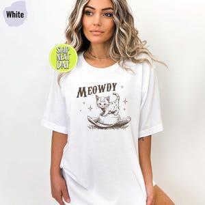 以下が含まれることがあります： 猫がカウボーイハットをかぶっているグラフィックと「MEOWDY」というテキストが書かれた白いTシャツです。
