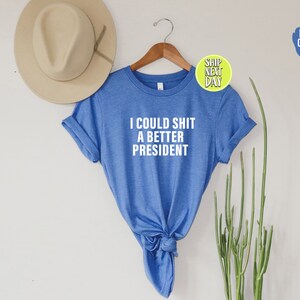 May include: A blue t-shirt with the text "I could shit a better president" printed in white on the front.