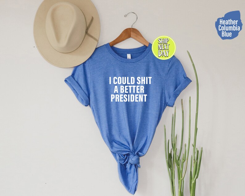 May include: A blue t-shirt with the text "I could shit a better president" printed in white on the front.
