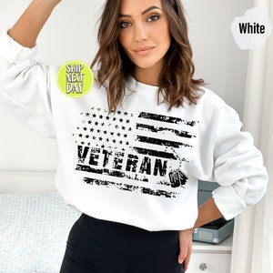 Peut inclure: Sweat-shirt blanc avec un motif de drapeau am&eacute;ricain d&eacute;lav&eacute; en noir et blanc et le mot "Veteran" imprim&eacute; dessus.