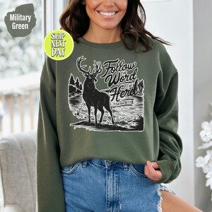 May include: Military green sweatshirt featuring a black silhouette of a deer with large antlers, standing in front of a lake and trees. The text "Follow the Word Not the Herd" and "Isaiah 8:11" are printed in white.