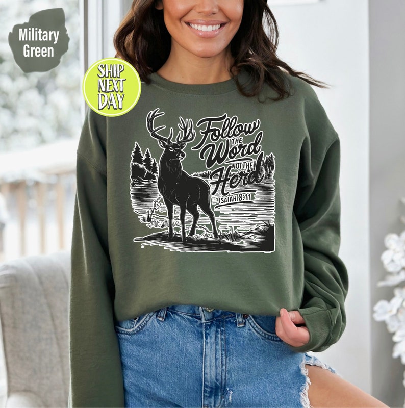 May include: Military green sweatshirt featuring a black silhouette of a deer with large antlers, standing in front of a lake and trees. The text "Follow the Word Not the Herd" and "Isaiah 8:11" are printed in white.