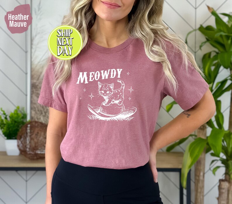 以下が含まれることがあります： ヘザーモーヴのTシャツに、カウボーイハットをかぶった猫の白いグラフィックと「MEOWDY」の文字がプリントされています。